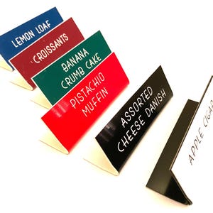May include: A set of six L-shaped food display signs in blue, maroon, green, red, black, and white. Each sign features white text reading Lemon Loaf, Croissants, Banana Crumb Cake, Pistachio Muffin, Assorted Cheese Danish, and Apple Cigar.