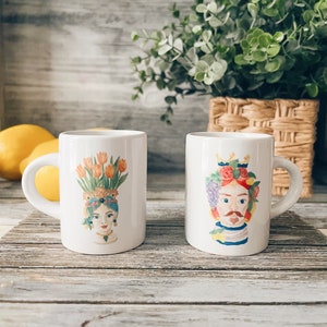 May include: Two white ceramic mugs with colorful illustrations of a woman and a man with fruit and flower crowns. The mugs are on a wooden surface.