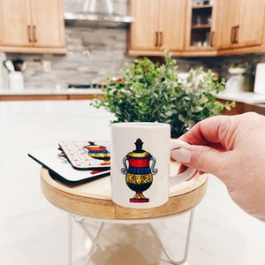 Op de afbeelding: Een wit keramiek espresso kopje met een kleurrijke illustratie van een vaas met een gezicht. Het kopje staat op een houten onderzetter op een witte metalen standaard.