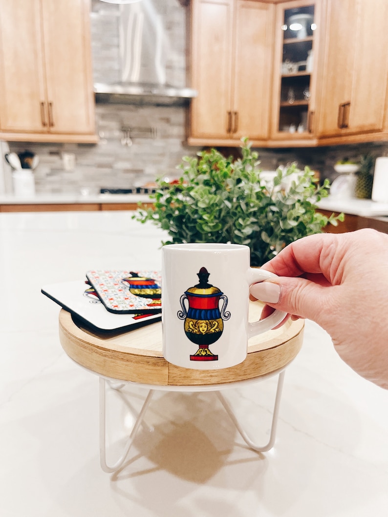 Op de afbeelding: Een wit keramiek espresso kopje met een kleurrijke illustratie van een vaas met een gezicht. Het kopje staat op een houten onderzetter op een witte metalen standaard.