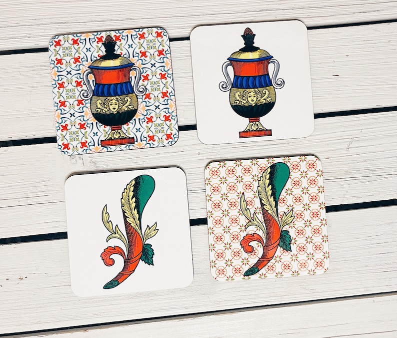 Op de afbeelding: Vier vierkante onderzetters met verschillende ontwerpen. De onderzetters zijn wit met een decoratief patroon. Een onderzetter heeft een rode, blauwe en gouden urn met een bloemmotief. Een andere onderzetter heeft een groen en rood gestileerd bloemmotief. De overige twee onderzetters hebben een herhalend patroon van kleine bloemen.