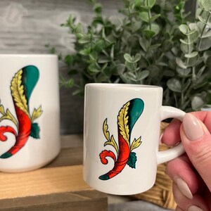 Op de afbeelding: Twee witte keramische mokken met een groen, rood en geel bloemenontwerp. De kleinere mok heeft een handvat.