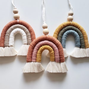 Könnte beinhalten: Drei bunte Makramee-Regenbogen-Wandbehänge mit Holzperlen. Jeder Regenbogen ist aus verschiedenen Garnfarben gefertigt und hat einen weißen Quastenfransen.