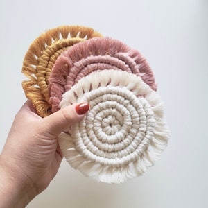 May include: Three round crocheted coasters with fringe. The top coaster is white, the middle coaster is pink, and the bottom coaster is yellow.