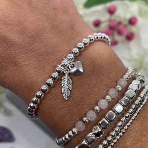 May include: A silver beaded bracelet with a heart and feather charm. The bracelet is stacked with other silver bracelets, including one with pink beads.