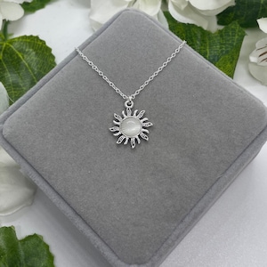 May include: A silver necklace with a sun pendant featuring a white opal stone. The necklace is displayed on a gray velvet box.