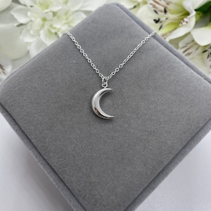 May include: A silver crescent moon pendant necklace on a silver chain. The necklace is displayed on a gray velvet box.