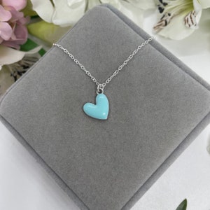 May include: A silver chain necklace with a light blue enamel heart pendant. The necklace is displayed on a gray velvet box.