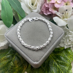 May include: A silver bracelet with an intricate, open-link design. The bracelet is displayed on a gray velvet surface, surrounded by white and pink flowers and green foliage. The bracelet has a lobster clasp closure.