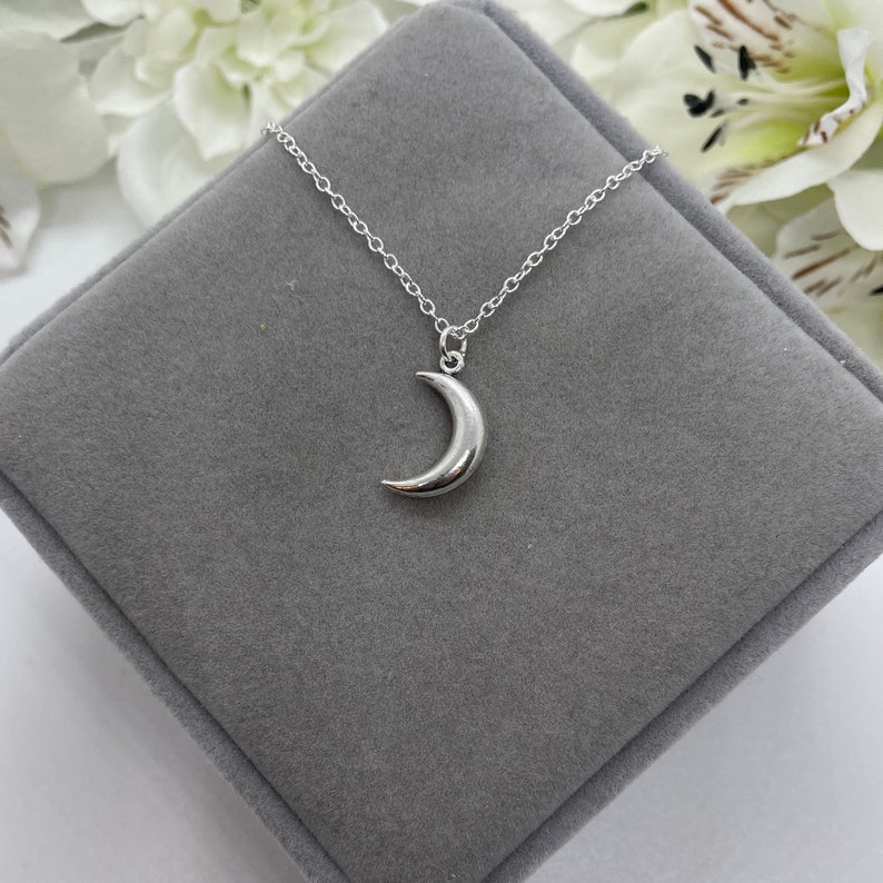May include: A silver crescent moon pendant necklace on a silver chain. The necklace is displayed on a gray velvet box.