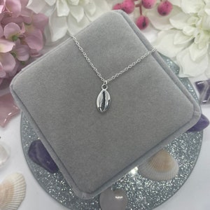 May include: A silver necklace with a small seashell pendant. The necklace is displayed in a grey velvet box.