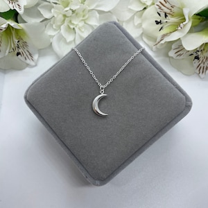 May include: A silver crescent moon pendant necklace on a silver chain, displayed on a gray velvet jewelry box.