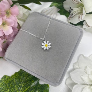 May include: A silver chain necklace with a white daisy pendant. The daisy has a yellow center and is on a gray velvet background.