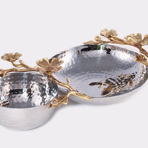 May include: A silver-toned, two-section serving bowl with a hammered texture. Gold-colored floral branch accents adorn the sides. The bowl is designed for serving snacks or appetizers, adding a touch of elegance to any table setting.