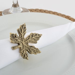 May include: Gold metal leaf napkin ring. The ring is in the shape of a maple leaf with a detailed design.