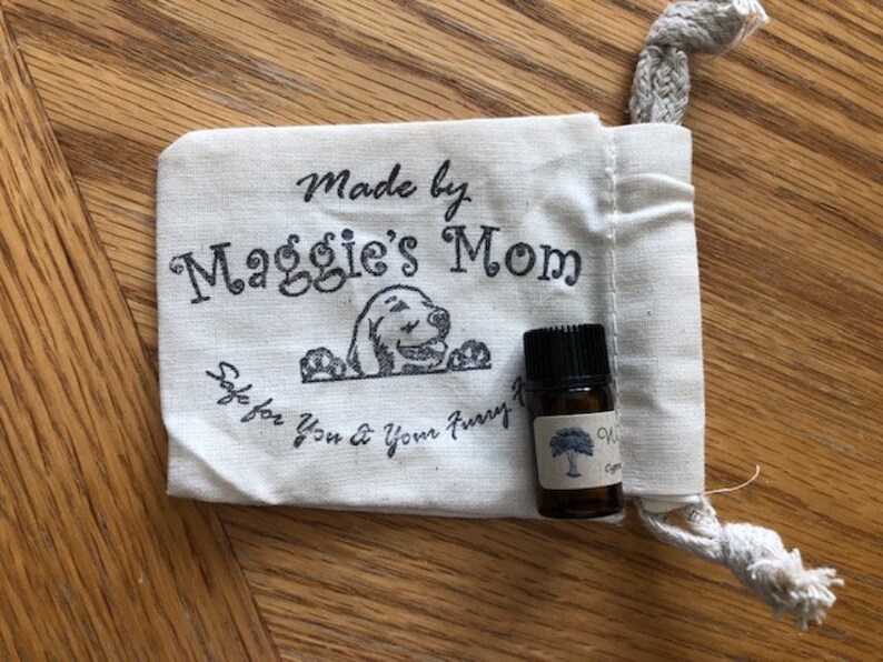 May include: A small white drawstring bag with the text "Made by Maggie's Mom Safe for you & your furry friend" and a cartoon dog. A small brown glass bottle with a white label and a tree design is sitting on the bag.
