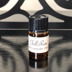 May include: A small brown glass bottle with a black cap. The bottle is labelled "Fall Rain" with the text "Sweet, Lavender, Clary" underneath.