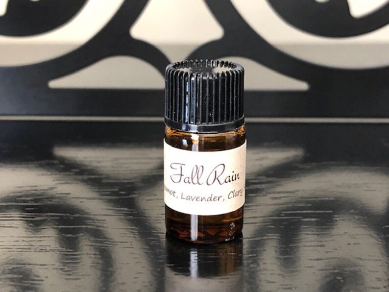 May include: A small brown glass bottle with a black cap. The bottle is labelled "Fall Rain" with the text "Sweet, Lavender, Clary" underneath.