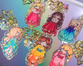 May include: Collection of acrylic keychains featuring cartoon characters in various outfits. Each keychain has a gold-colored star-shaped clasp with a crystal accent. The characters are depicted in vibrant colors, with pink, yellow, and blue dresses.