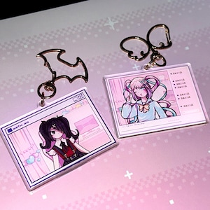May include: Two acrylic keychains featuring anime-style illustrations. One keychain has a bat-shaped charm, the other has a double ring charm. The illustrations depict characters in a pink-themed interface, with the word "webcam" visible on one.