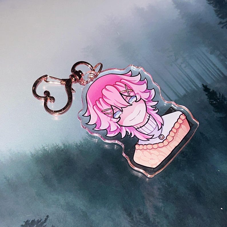 May include: A clear acrylic keychain featuring a cartoon character with pink hair and blue eyes. The character is wearing a white sweater with a tan top. The keychain has a rose gold clasp.
