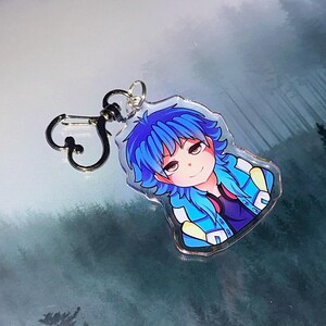 May include: A clear acrylic keychain featuring a cartoon character with blue hair and a blue jacket. The character has a smiling expression. The keychain has a silver-colored clasp.
