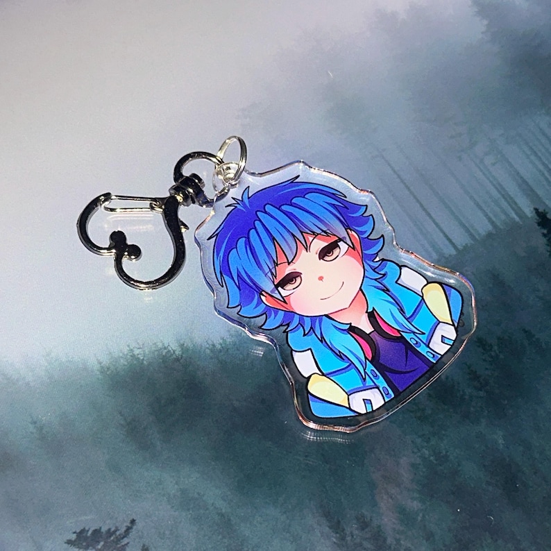 May include: A clear acrylic keychain featuring a cartoon character with blue hair and a blue jacket. The character has a smiling expression. The keychain has a silver-colored clasp.