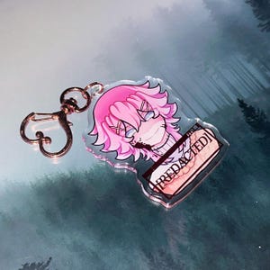 May include: A clear acrylic keychain featuring a cartoon character with pink hair and blue eyes. The keychain has a rose gold clasp and a rectangular tag that reads "REDACTED".