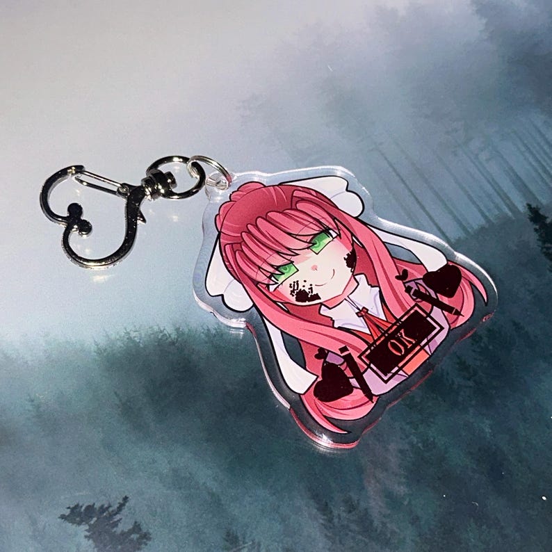 May include: A clear acrylic keychain featuring a cartoon character with pink hair and green eyes. The character has black markings on their face and holds a sign that says "OK". The keychain has a silver-colored clasp.
