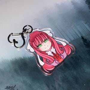 May include: A clear acrylic keychain featuring a cartoon illustration of a character with pink hair, green eyes, and a red bow. The keychain has a silver-colored clasp for attachment.