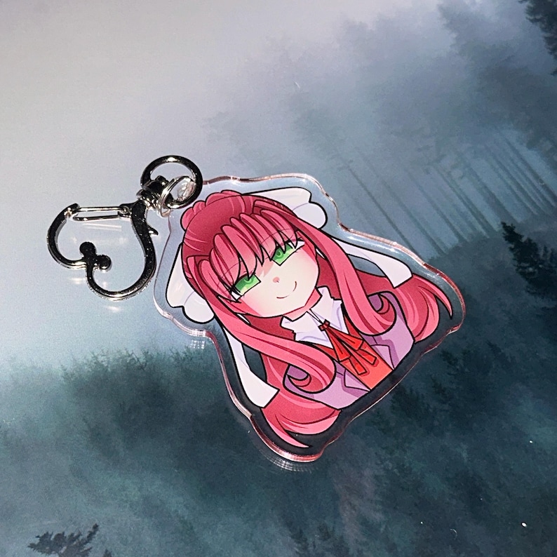 May include: A clear acrylic keychain featuring a cartoon illustration of a character with pink hair, green eyes, and a red bow. The keychain has a silver-colored clasp for attachment.