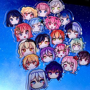 Puede incluir: Una colección de charms de acrílico de personajes de anime de colores vivos, con diferentes personajes femeninos con diferentes peinados y expresiones. Los charms están dispuestos en un grupo sobre un fondo azul.