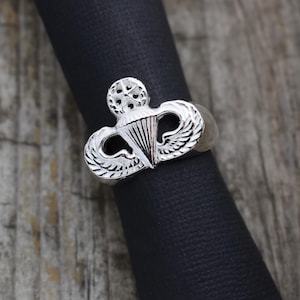 May include: A silver ring with a detailed design of a paratrooper badge. The ring is on a black surface.