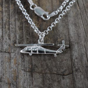 May include: A silver-colored helicopter pendant hangs from a delicate chain necklace. The pendant is a detailed miniature of a helicopter, with visible rotors and tail. The necklace is displayed against a rustic wooden background.