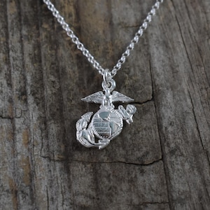 May include: A silver-colored necklace featuring a pendant with the United States Marine Corps emblem. The emblem includes an eagle, globe, and anchor design. The necklace is displayed on a weathered wooden surface.