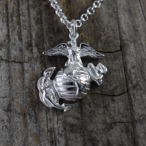 May include: Silver pendant necklace featuring the United States Marine Corps emblem. The emblem depicts a globe with an eagle perched on top and an anchor on the side.