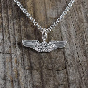 May include: Silver-toned pendant necklace featuring a detailed wing design. The pendant is suspended from a delicate chain, set against a rustic wooden background. The wings are intricately detailed, showcasing a classic aviation theme.