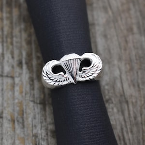 May include: A silver ring with a detailed design of a parachute and wings. The ring is on a black velvet background.