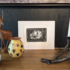 May include: A black and white print of a television set with a distorted image inside. The print is framed in a white frame and is sitting on a wooden table with a wooden deer figurine, a yellow vase with a black and white animal print, and a metal candle holder with a white candle.