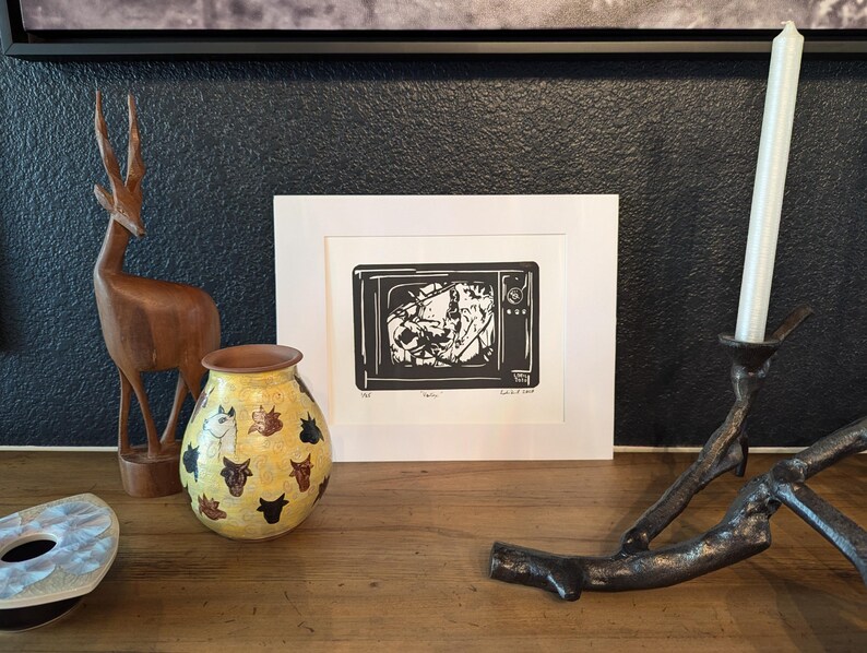 May include: A black and white print of a television set with a distorted image inside. The print is framed in a white frame and is sitting on a wooden table with a wooden deer figurine, a yellow vase with a black and white animal print, and a metal candle holder with a white candle.