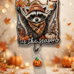 May include: A glittery badge reel featuring a raccoon wearing a beanie, heart-shaped sunglasses, and a plaid scarf. The raccoon holds a maple leaf and a coffee cup. The text 'tis the season' is printed below, with a small orange pumpkin charm attached.