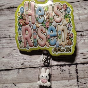 May include: A badge reel featuring a glittery lime green border and the text He Is Risen with Matthew 28:6. The design includes pastel-coloured letters, small rabbits, flowers, and Easter eggs. A small white rabbit charm with a pink bow is attached.