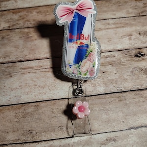 Pu&ograve; includere: Un badge reel Red Bull con un fiocco rosa e accenti floreali. Il badge reel presenta un cinturino in plastica trasparente e un ciondolo a forma di fiore rosa. Il design della lattina Red Bull &egrave; su uno sfondo glitterato argento.