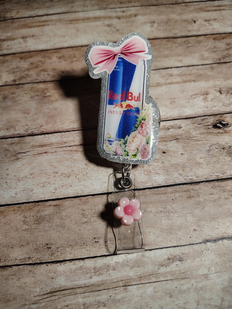 Pu&ograve; includere: Un badge reel Red Bull con un fiocco rosa e accenti floreali. Il badge reel presenta un cinturino in plastica trasparente e un ciondolo a forma di fiore rosa. Il design della lattina Red Bull &egrave; su uno sfondo glitterato argento.