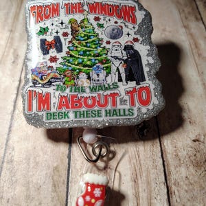 May include: A badge reel with a festive design featuring Star Wars characters and a Christmas tree. The text reads "From the windows to the walls I'm about to deck these halls." The reel has a red and white Christmas stocking charm.