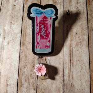 May include: A retractable badge reel featuring a pink and white Alani Nu Energy drink can design with a blue bow. The reel has a black border and a pink flower accent.