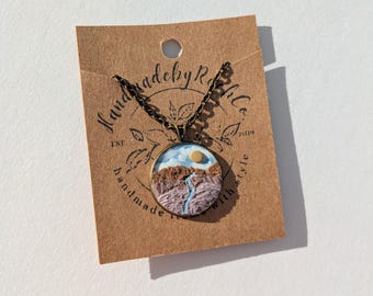 May include: A round, brown, and blue necklace pendant with a painted landscape scene. The pendant is on a brown card with the text "Handmade by Rille" and "Est. 2019".