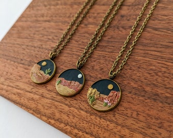 May include: Three bronze-toned pendant necklaces with desert landscape designs. Each pendant features a miniature scene with a night sky, moon, and desert elements like cacti and rock formations. The necklaces rest on a wooden surface.