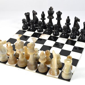 Puede incluir: Un tablero de ajedrez de mármol blanco y negro con un juego de piezas de ajedrez blancas y negras. Las piezas blancas están hechas de hueso y las piezas negras están hechas de madera oscura.