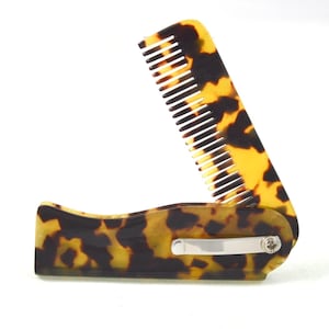 Könnte beinhalten: Ein Klappkamm mit Schildpattmuster. Der Kamm ist geöffnet und zeigt weiße Zähne. Das Scharnier und der Verschluss sind silberfarben. Der Kamm ist vor einem weißen Hintergrund platziert.